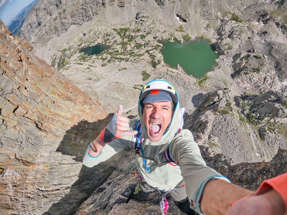 Pascal high on the Petit Grepon, shouting and throwing a shaka, two green lakes far below